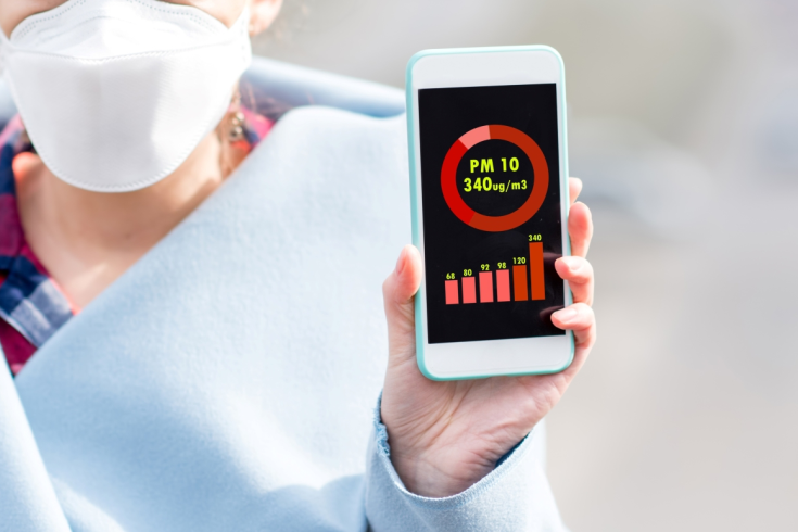 Osoba w maseczce ochronnej pokazująca telefon z aplikacją wskazującą bardzo wysokie stężenie pyłu PM10 w powietrzu (340 ug/m³)