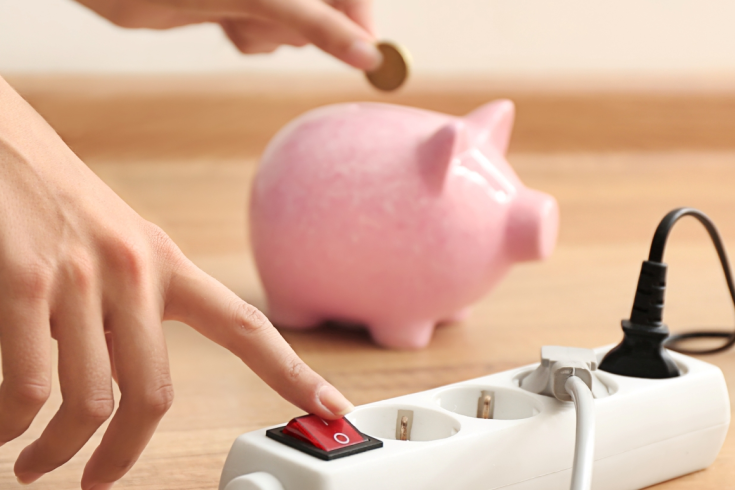 Osoba naciska wyłącznik na listwie zasilającej, podczas gdy w tle wrzuca monetę do różowej skarbonki