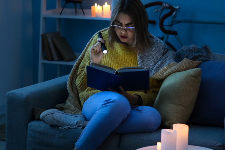 Kobieta siedząca na kanapie w ciemnym pokoju, owinięta kocem, czyta książkę przy latarce podczas przerwy w dostawie prądu. W tle palą się świece.