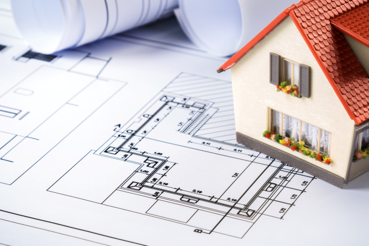 Makieta domu jednorodzinnego z czerwonym dachem ustawiona na rozłożonych planach architektonicznych, w tle zwinięte projekty techniczne